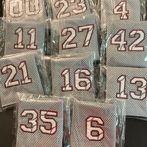 May include: A collection of baseball jerseys in clear plastic packaging. Each jersey features a white number with baseball stitching on a maroon background. The jerseys are gray with a mesh design.