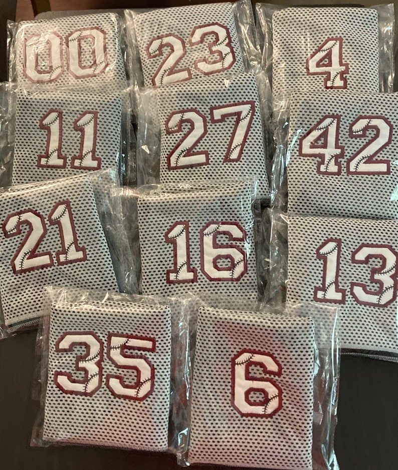 May include: A collection of baseball jerseys in clear plastic packaging. Each jersey features a white number with baseball stitching on a maroon background. The jerseys are gray with a mesh design.