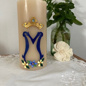 Op de afbeelding: Een beige kaars met een blauw en gouden geschilderd ontwerp. De kaars heeft een kroon met een blauwe edelsteen, een blauwe letter "M" en gele en blauwe bloemen.