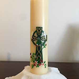 May include: A white candle with a green Celtic cross and red and white flowers. The candle is on a white cloth.