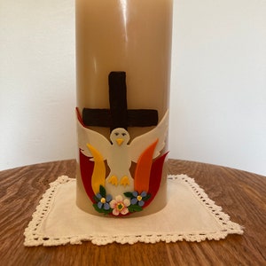 May include: A beige pillar candle with a decorative design featuring a dark brown cross, a white dove, and colorful accents. The candle sits on a white lace doily on a wooden table.