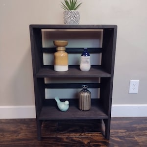 May include: A black wooden shelf unit with three shelves. The top shelf has a potted plant with green leaves. The middle shelf has a yellow and white ceramic vase and a blue and white striped vase. The bottom shelf has a blue bird figurine and a black and white striped vase.