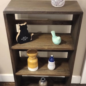 May include: A dark brown wooden shelf unit with three shelves. The top shelf has a small potted plant with green leaves. The second shelf has a black cat figurine and a wooden sign that says "Life is better with a cat". The third shelf has a blue and white striped vase, a yellow and white vase, and a turquoise bird figurine.