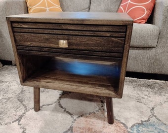 May include: A dark brown wooden end table with a single drawer and a lower shelf. The drawer has a gold metal pull. The table has a distressed finish and sits on four short legs.