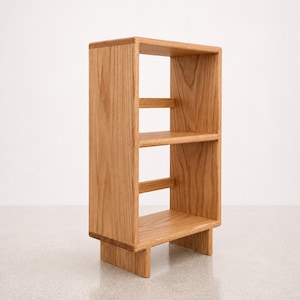 May include: A small, light brown wooden bookshelf with two shelves. The bookshelf is rectangular with a simple, modern design. It has a sturdy base and is made of natural wood.