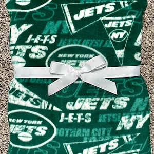 Puede incluir: Una manta de franela verde con una cinta blanca atada alrededor. La manta está cubierta con un patrón del logotipo de los New York Jets, las palabras "Jets" y "New York".