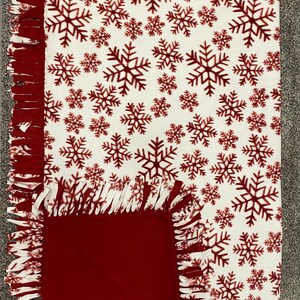 May include: A cozy, rectangular fleece blanket with a white background and a pattern of burgundy snowflakes. The blanket has a solid burgundy square on one corner and a fringed edge.