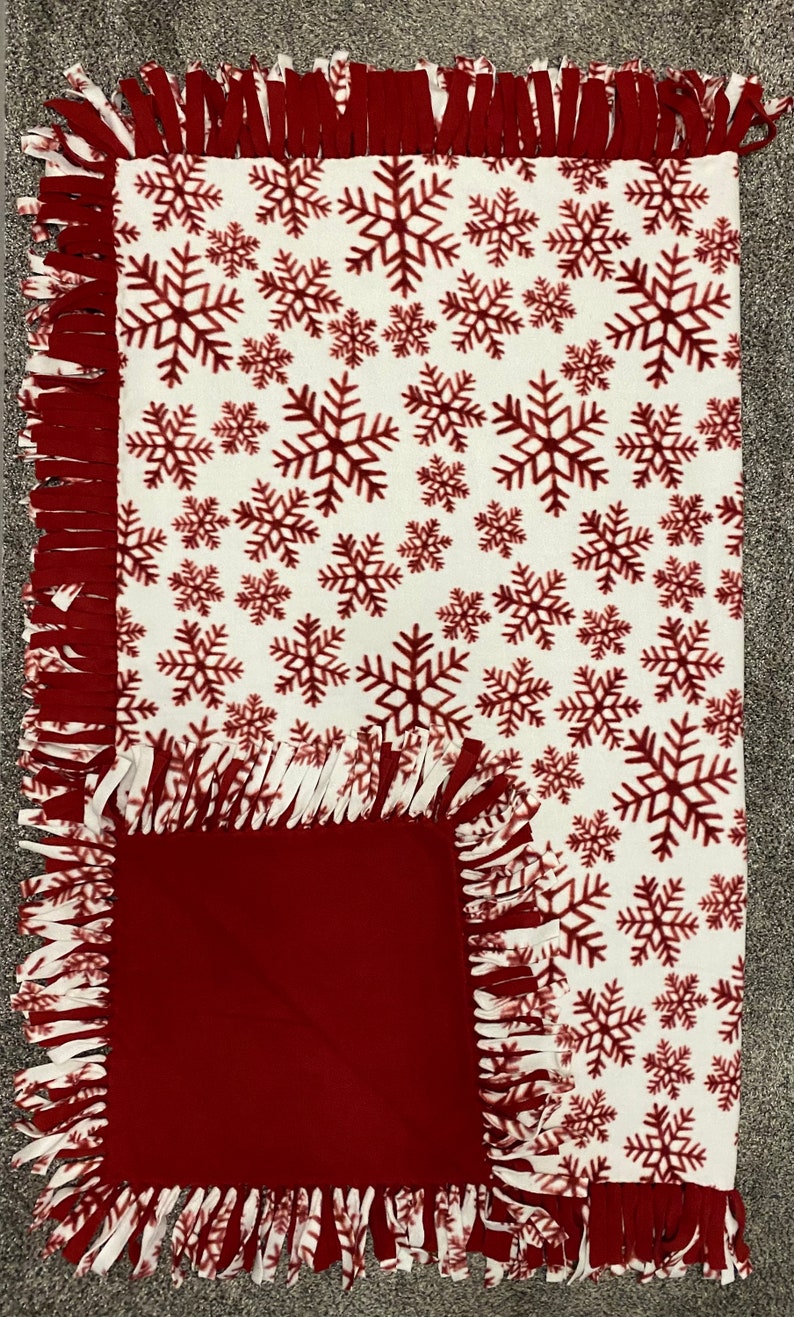 May include: A cozy, rectangular fleece blanket with a white background and a pattern of burgundy snowflakes. The blanket has a solid burgundy square on one corner and a fringed edge.