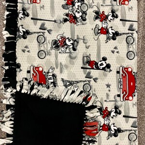 May include: A gray and white fleece blanket with a black fleece backing. The blanket features a repeating pattern of Mickey Mouse and Minnie Mouse in red, black, and white. The pattern includes hearts, stars, and Eiffel Towers. The blanket has a fringe border.
