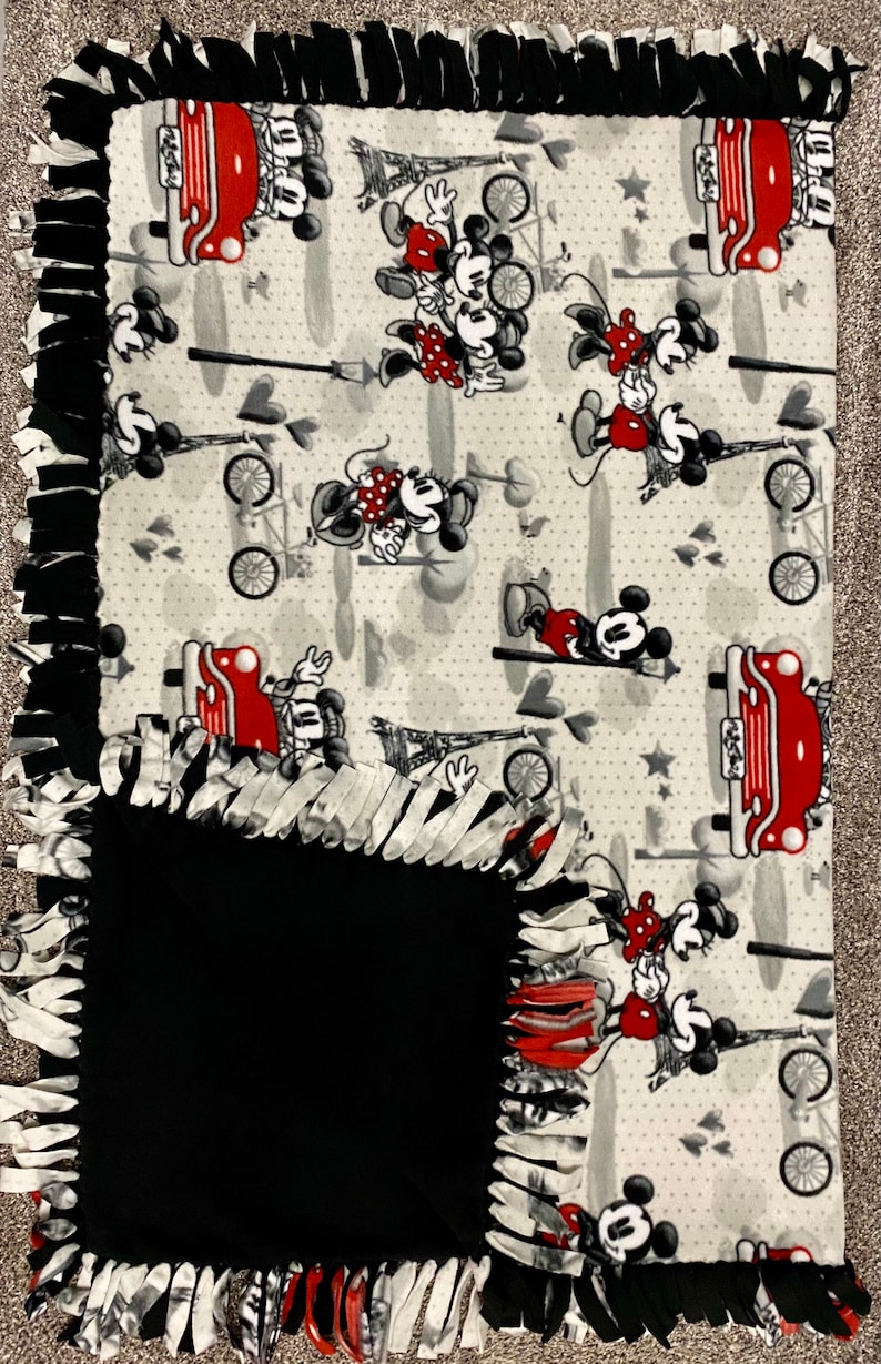 May include: A gray and white fleece blanket with a black fleece backing. The blanket features a repeating pattern of Mickey Mouse and Minnie Mouse in red, black, and white. The pattern includes hearts, stars, and Eiffel Towers. The blanket has a fringe border.