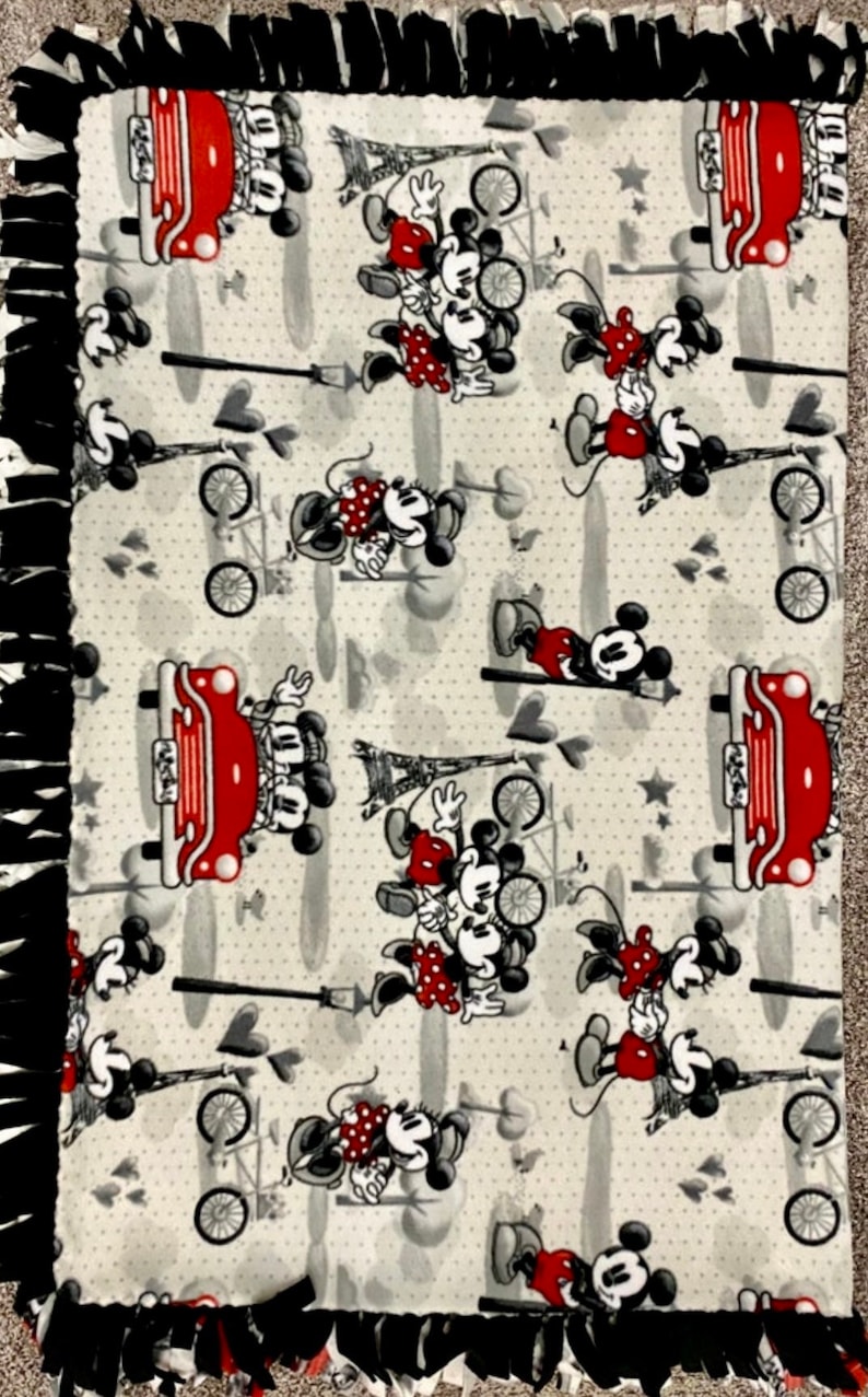 May include: A white fleece blanket with a black and red fringe border. The blanket features a repeating pattern of Mickey Mouse and Minnie Mouse in various poses, including riding in a red car, riding a bicycle, and standing together. The background is a light gray with white polka dots.