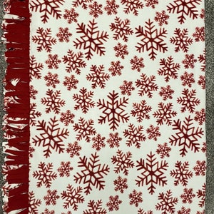 May include: A white fleece blanket with a repeating pattern of burgundy snowflakes. The edges are finished with a burgundy fringe. This cozy blanket is perfect for winter or holiday decor.