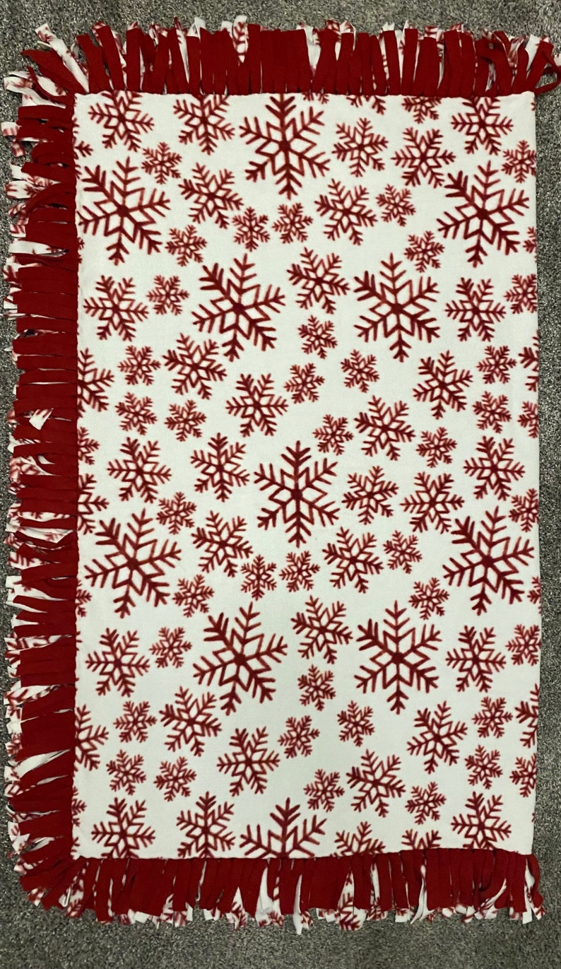 May include: A white fleece blanket with a repeating pattern of burgundy snowflakes. The edges are finished with a burgundy fringe. This cozy blanket is perfect for winter or holiday decor.