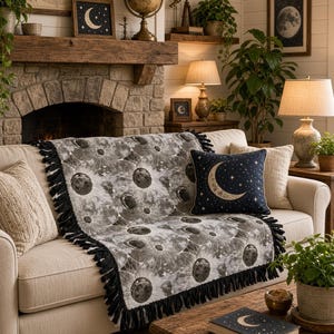 May include: A gray and white throw blanket featuring a repeating moon phase pattern with black fringe edges, paired with a square navy blue throw pillow displaying a gold crescent moon and star design on a light-colored sofa.