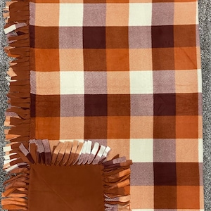 May include: A brown, orange and white plaid fleece blanket with a brown fleece square in the centre. The blanket has a fringe border.