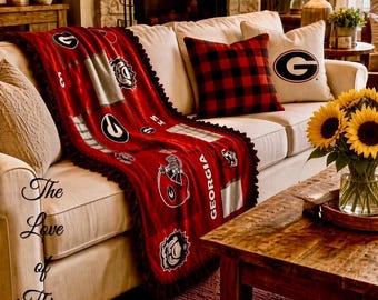 May include: A red and black fleece throw blanket featuring the Georgia Bulldogs logo, football helmet graphics, and the word GEORGIA. The blanket has a dark fringe border and is draped over a cream-colored sofa with a red and black plaid pillow.