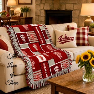 Op de afbeelding: Een fleece deken met een patchwork ontwerp van karmozijnrode en grijze vierkanten met Indiana University logo's en tekst. De deken heeft een geknoopte franjerand en ligt gedrapeerd over een crèmekleurige bank met bijpassende sierkussens.