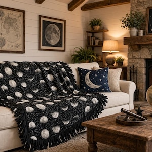 May include: A black fleece throw blanket featuring a repeating pattern of white moon phases and small stars, finished with a fringed edge. The blanket is draped over a light-colored sofa, accompanied by a dark blue square pillow with a crescent moon.