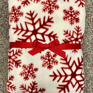 May include: A folded white fleece blanket with a repeating pattern of large, dark red snowflakes. The blanket is tied with a red ribbon and bow, suggesting it's a gift. The blanket is soft and cozy, perfect for winter.