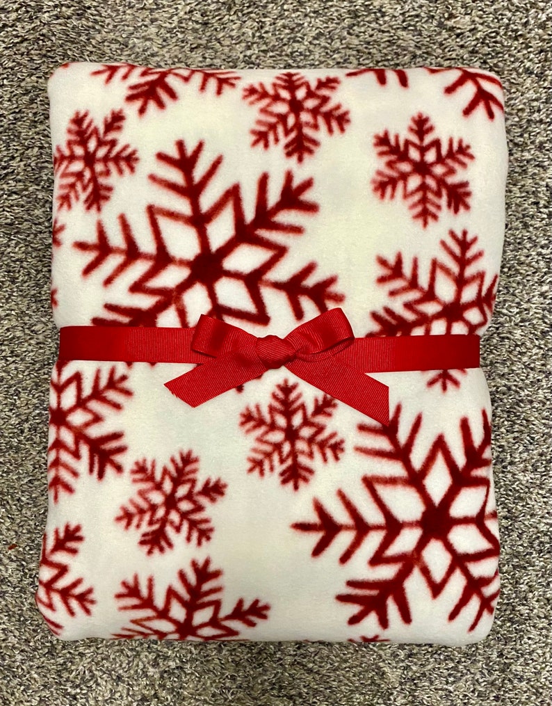 May include: A folded white fleece blanket with a repeating pattern of large, dark red snowflakes. The blanket is tied with a red ribbon and bow, suggesting it's a gift. The blanket is soft and cozy, perfect for winter.