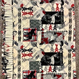 May include: A white fleece blanket with a patchwork design featuring black and gray deer, pine cones, and red and white geometric patterns. The blanket has a white fringe border.