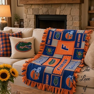 Könnte beinhalten: Eine Fleecedecke mit einem Patchwork-Design aus blauen und orangefarbenen Quadraten, verziert mit Florida Gators-Logos, Football-Helmen sowie dem Schriftzug THE SWAMP, FLORIDA und Gators. Die Decke hat einen Rand mit geknoteten Fransen.