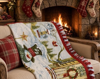 May include: A rectangular throw blanket featuring a winter scene with a snowman, a house with a wreath on the door, and a red cardinal. The blanket has a white base with a red plaid ruffled border and is draped over a beige upholstered sofa.