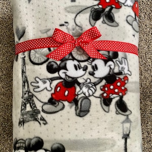 May include: A gray fleece blanket with a white polka dot pattern featuring Mickey and Minnie Mouse in a Parisian setting. The blanket is tied with a red polka dot ribbon.