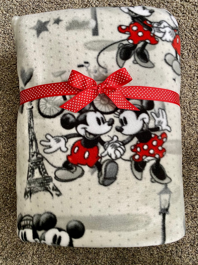 May include: A gray fleece blanket with a white polka dot pattern featuring Mickey and Minnie Mouse in a Parisian setting. The blanket is tied with a red polka dot ribbon.