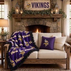 May include: A purple fleece throw blanket with a black fringed edge, featuring a repeating pattern of the Minnesota Vikings logo and the text VIKINGS in yellow and white. A matching purple square throw pillow with the team logo is also present.