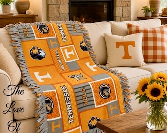 May include: A fleece throw blanket featuring a patchwork design in orange, white, and grey. The pattern includes the Tennessee Volunteers logo, the text "VOLS," "TENNESSEE," and "VOLUNTEERS," along with a mascot graphic and a large letter T.