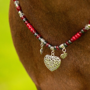 May include: A beaded necklace with red, silver, and black beads. The necklace features three silver heart charms, with the largest heart having a filigree design. The necklace is displayed on a brown surface against a blurred green background.