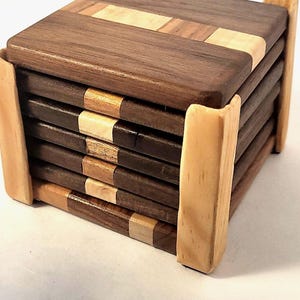 Puede incluir: Un juego de seis posavasos cuadrados de madera con un patrón de vetas de madera marrón oscuro y claro. Los posavasos se guardan en un soporte de madera clara. Cada posavasos mide aproximadamente 10 cm cuadrados.
