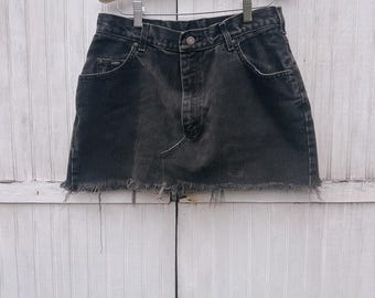 May include: A black denim skirt with a frayed hem hangs from a wooden hanger. The skirt has a classic five-pocket design and a button closure. The denim appears faded, giving it a vintage look. The skirt is displayed against a white, paneled background.