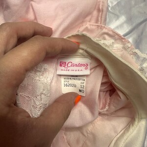 Puede incluir: Primer plano de una prenda de beb&eacute; rosa con ribete de encaje blanco. La prenda tiene una etiqueta que dice "Carter's MADE IN U.S.A." con detalles sobre la tela y la talla 12 MO.