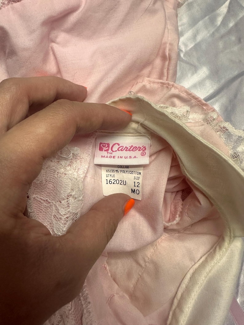 Puede incluir: Primer plano de una prenda de beb&eacute; rosa con ribete de encaje blanco. La prenda tiene una etiqueta que dice "Carter's MADE IN U.S.A." con detalles sobre la tela y la talla 12 MO.