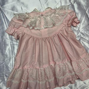 Puede incluir: Vestido rosa p&aacute;lido con cuello y borde de encaje blanco. El vestido tiene mangas cortas y cierre de botones en la espalda. La tela parece ligera, posiblemente de algod&oacute;n. El vestido tiene una est&eacute;tica vintage.
