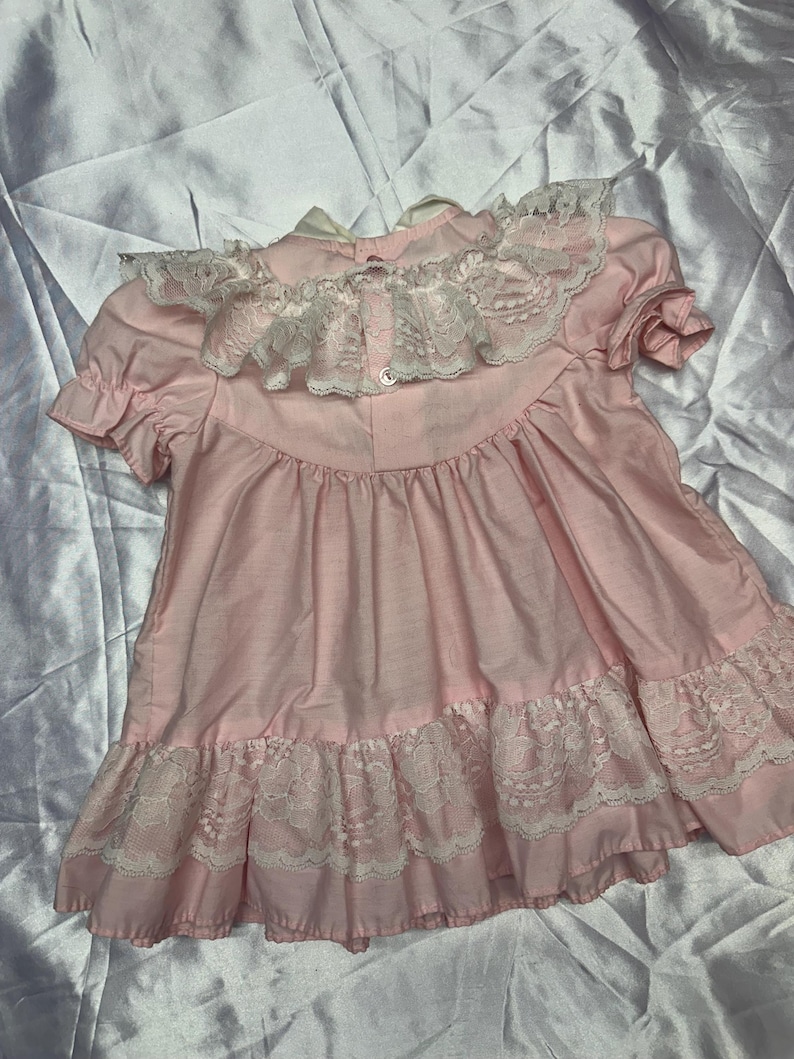 Puede incluir: Vestido rosa p&aacute;lido con cuello y borde de encaje blanco. El vestido tiene mangas cortas y cierre de botones en la espalda. La tela parece ligera, posiblemente de algod&oacute;n. El vestido tiene una est&eacute;tica vintage.