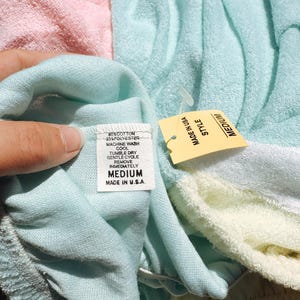 May include: A medium-sized, pastel-colored terry cloth garment with a tag that reads "MADE IN U.S.A." and "MEDIUM". The garment is a blend of cotton and polyester. The tag also includes washing instructions.
