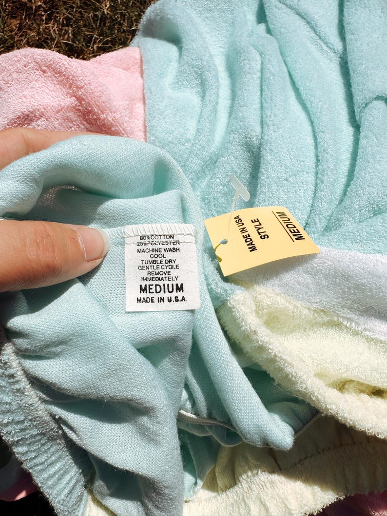 May include: A medium-sized, pastel-colored terry cloth garment with a tag that reads "MADE IN U.S.A." and "MEDIUM". The garment is a blend of cotton and polyester. The tag also includes washing instructions.