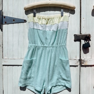 May include: A pastel-colored terry cloth romper with a strapless design. The romper features a yellow, white, and light blue striped top, an elastic waist, and two front pockets. It hangs on a wooden hanger against a white wooden door.