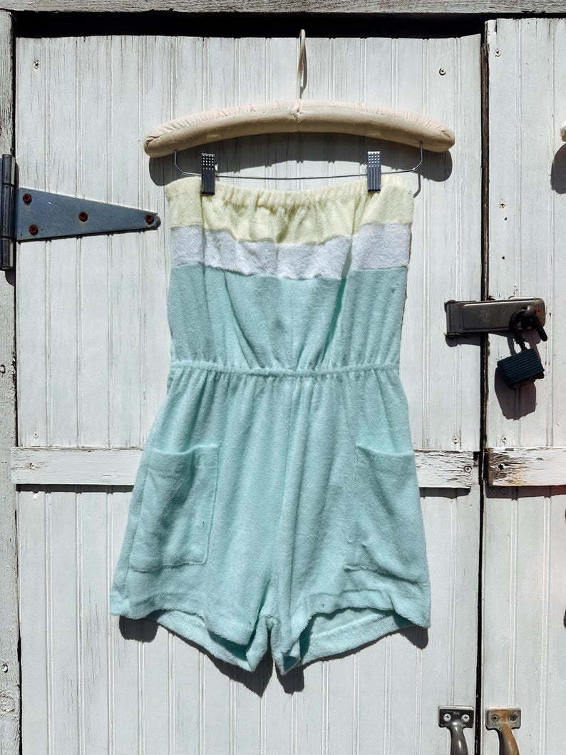 May include: A pastel-colored terry cloth romper with a strapless design. The romper features a yellow, white, and light blue striped top, an elastic waist, and two front pockets. It hangs on a wooden hanger against a white wooden door.
