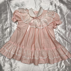 Puede incluir: Vestido rosa claro con cuello blanco y ribete de encaje. El vestido presenta mangas cortas, un dobladillo con volantes y una cinta en el escote. La prenda est&aacute; hecha de una tela suave.