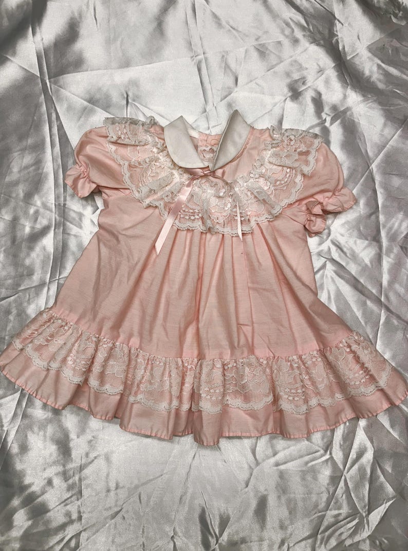 Puede incluir: Vestido rosa claro con cuello blanco y ribete de encaje. El vestido presenta mangas cortas, un dobladillo con volantes y una cinta en el escote. La prenda est&aacute; hecha de una tela suave.