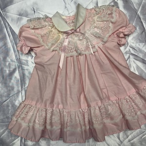 Puede incluir: Un vestido rosa vintage con cuello blanco y ribete de encaje. El vestido presenta mangas cortas, un dobladillo con volantes y un lazo de cinta de sat&eacute;n. El vestido es probablemente para un ni&ntilde;o.