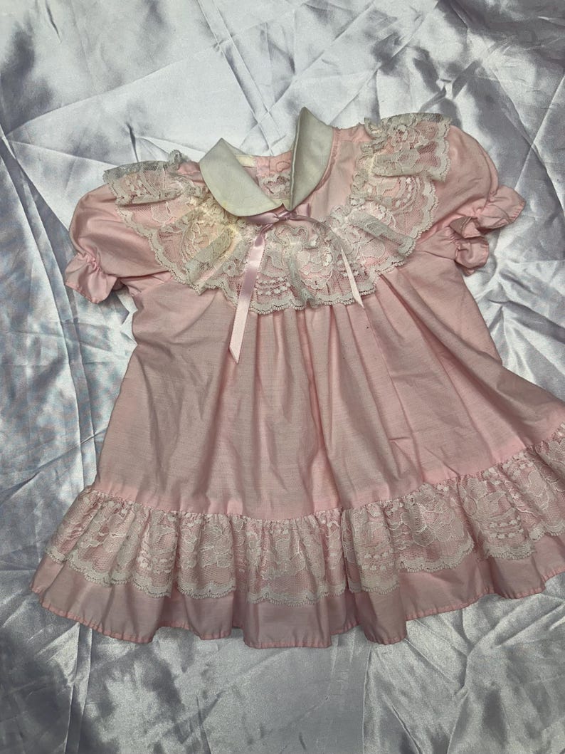 Puede incluir: Un vestido rosa vintage con cuello blanco y ribete de encaje. El vestido presenta mangas cortas, un dobladillo con volantes y un lazo de cinta de sat&eacute;n. El vestido es probablemente para un ni&ntilde;o.