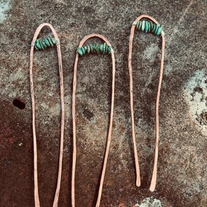 May include: Three copper hair pins with a U-shaped top adorned with small, turquoise-colored stones. The pins have a hammered texture and are arranged against a mottled, rusty-brown background, creating a rustic aesthetic. The pins are of varying lengths.