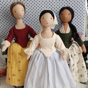 May include: Three handmade fabric dolls dressed in historical attire. The dolls have stitched facial features and wear dresses in shades of red, yellow, green, and blue. The dresses feature lace trim and floral or striped patterns, evoking a vintage aesthetic.
