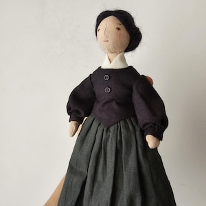 May include: A handmade cloth doll with a light skin tone, black hair, and a black dress with a white collar. The doll's dress has two buttons and a grey skirt. The doll is a soft sculpture.