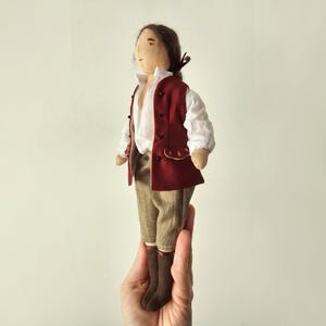 May include: A handmade doll in historical clothing. The doll is dressed in a white shirt, a burgundy vest, tan trousers, and brown boots. The doll has brown hair tied back. The doll is held in a hand.